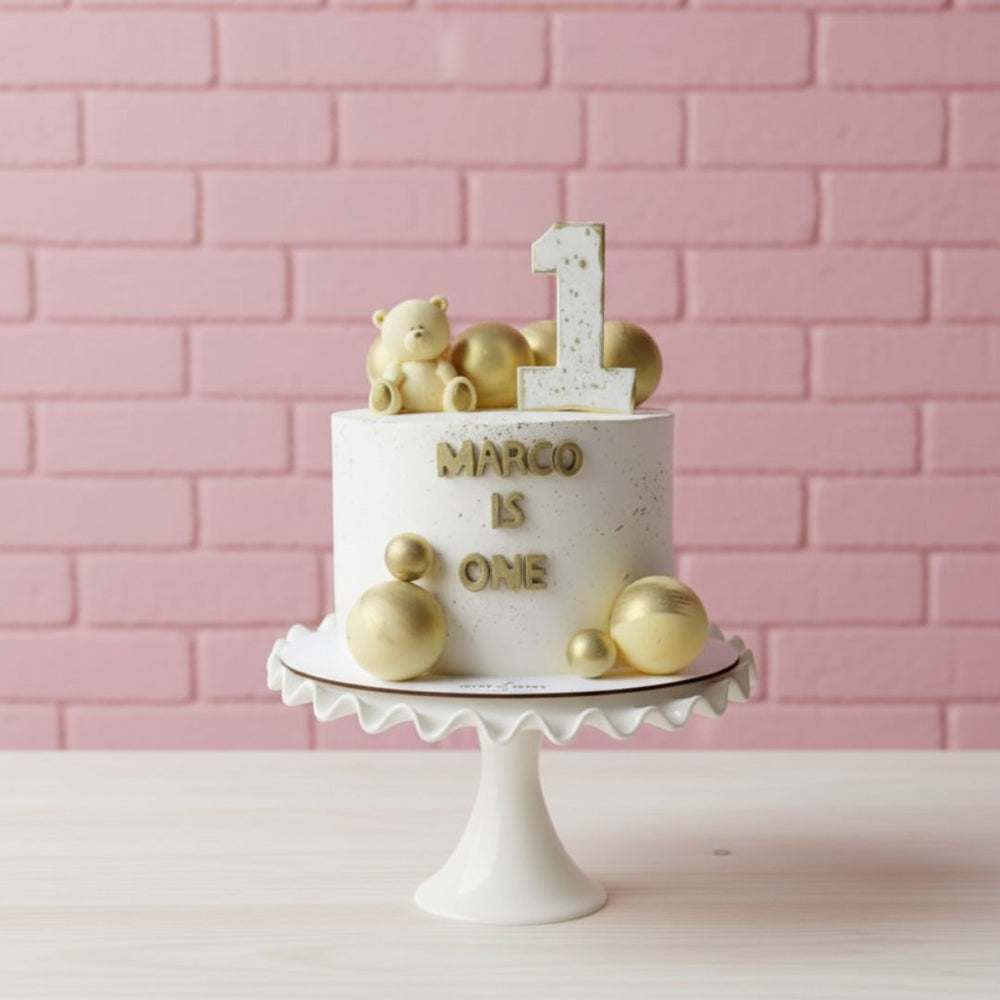 White cake with gold decorations and 'Marco is one' text on a white stand against a pink brick wall.
