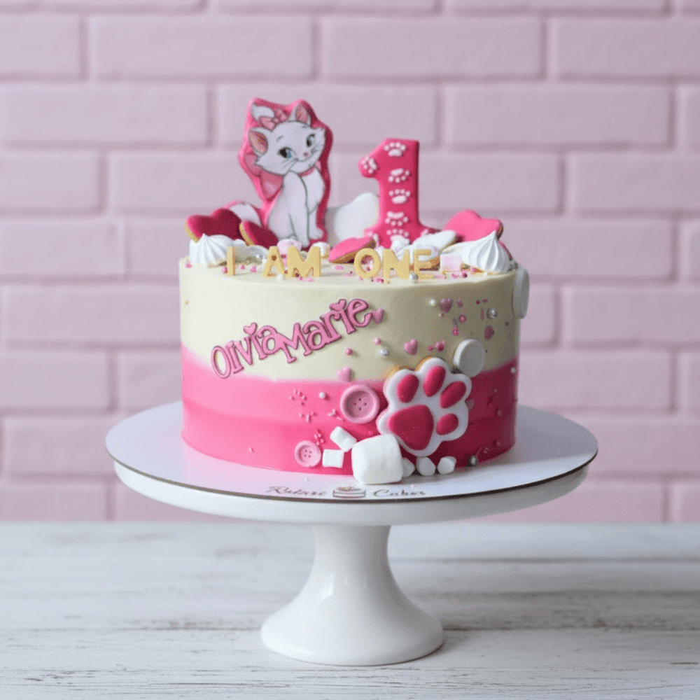 Pink and white birthday cake with cat-themed decorations on a white stand against a pink brick wall.