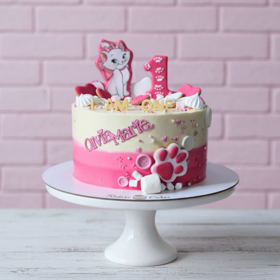 Pink and white birthday cake with cat-themed decorations on a white stand against a pink brick wall.