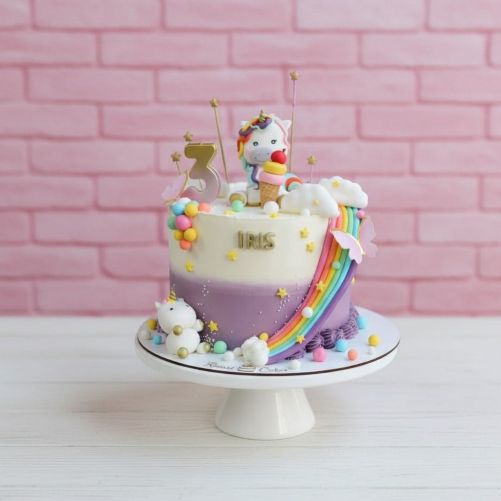 Colorful unicorn-themed birthday cake with rainbow and stars on a white stand against a pink brick wall.