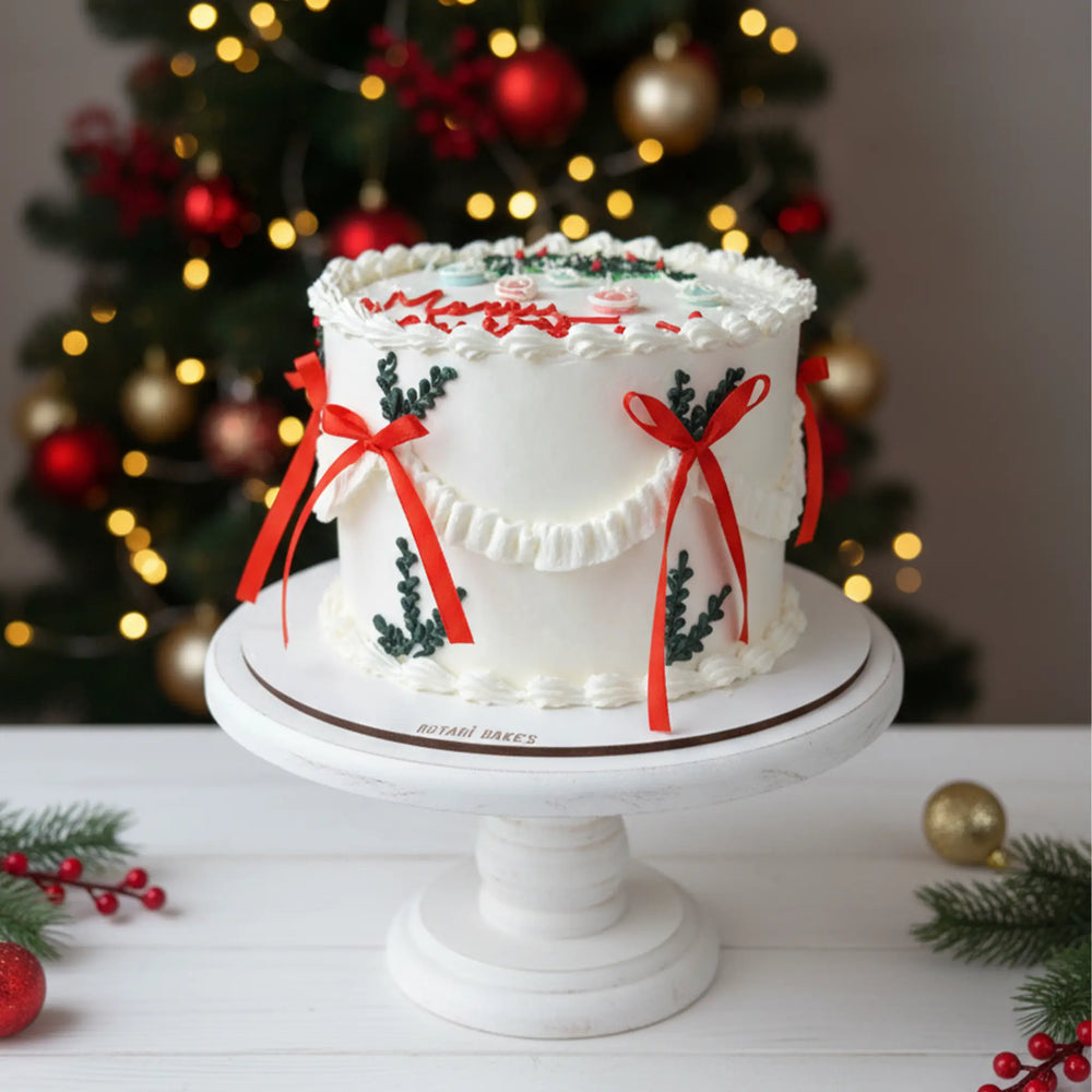 Christmas cake with red ribbons and green holly leaves on a white stand in front of a Christmas tree.