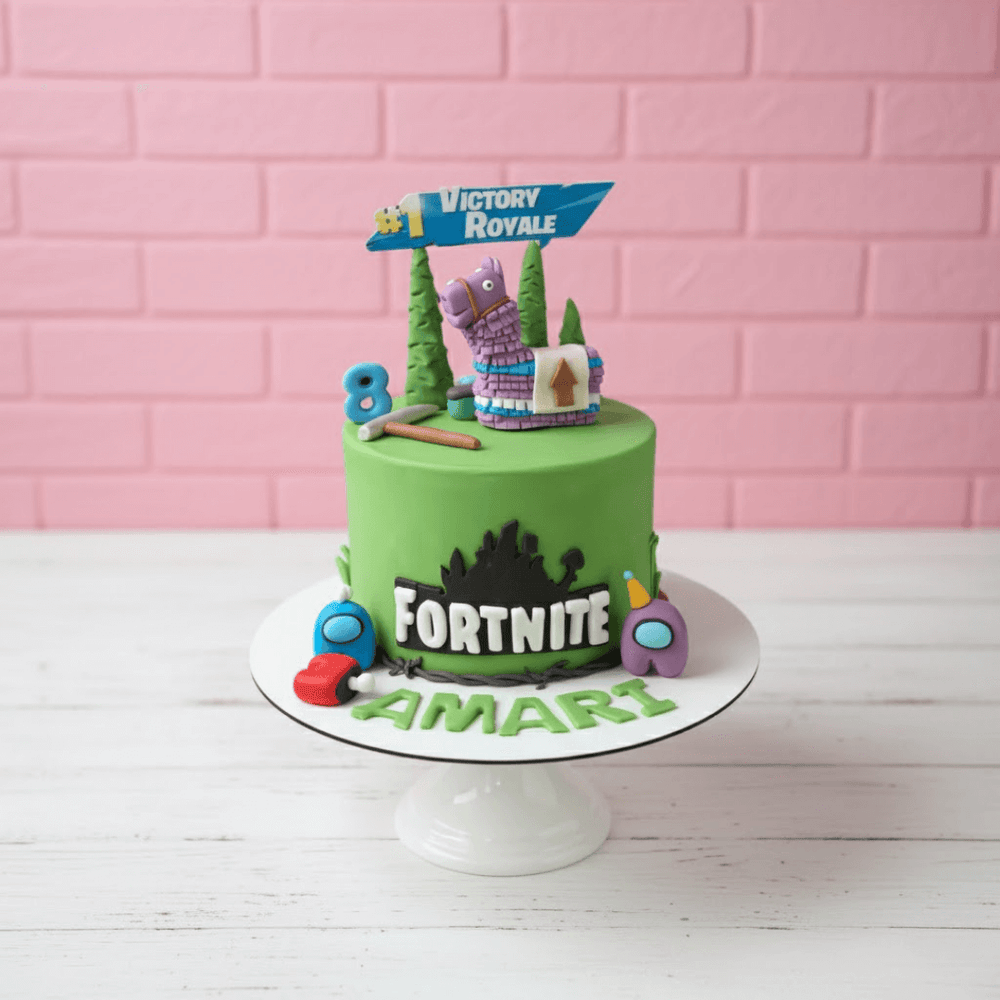 Green Fortnite cake with decorative elements on a white stand against a pink brick wall.
