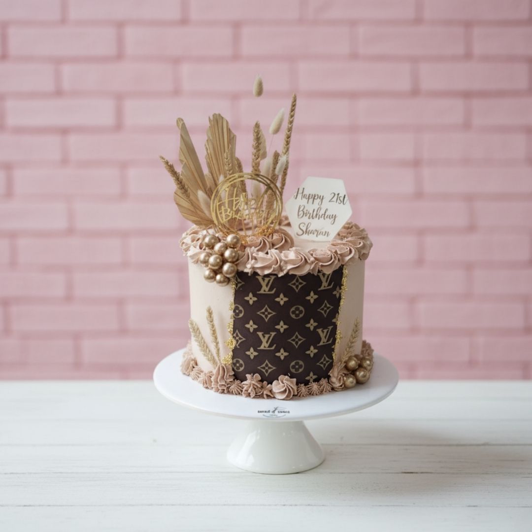 Decorative LV cake with gold accents on a white stand against a pink brick wall.