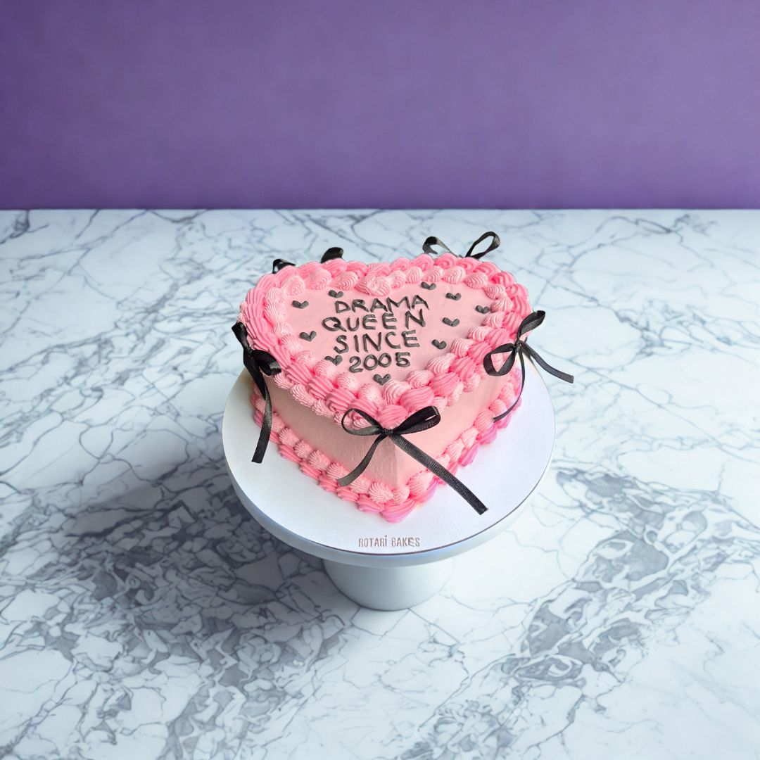 Heart-shaped pink cake with text drama queen since 2005 on it.