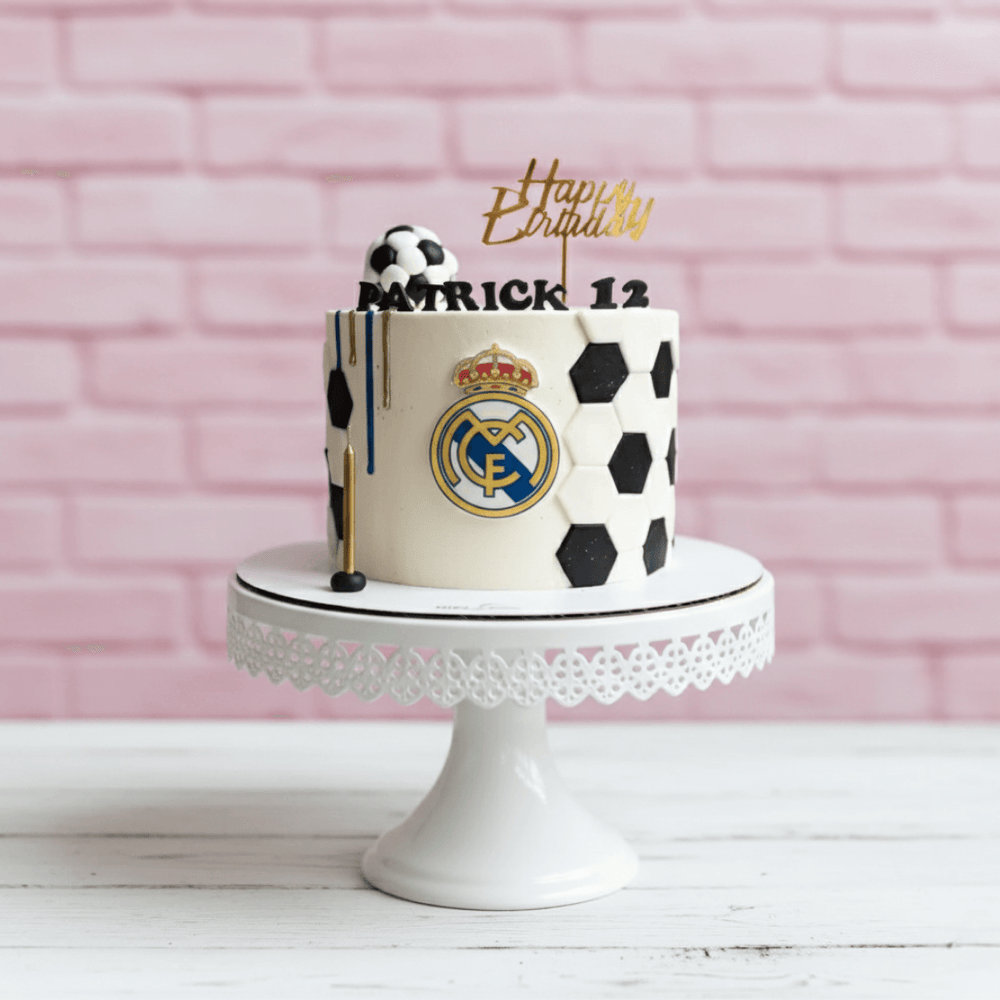 Real madrid theme Birthday cake with Real Madrid logo on a white stand against a pink brick wall.