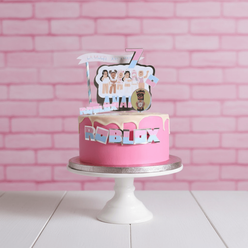Pink cake with Roblox-themed decorations on a white stand against a pink brick wall.