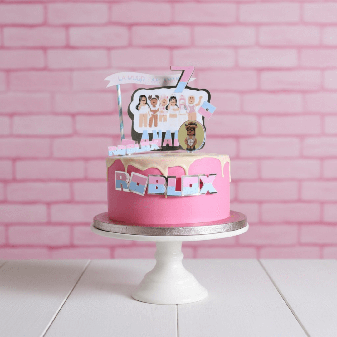 Pink cake with Roblox-themed decorations on a white stand against a pink brick wall.