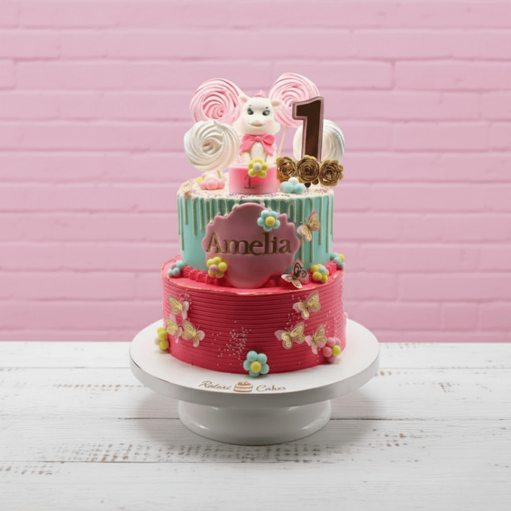 Two-tier aristocat theme birthday cake with pink and blue colors, decorated with a teddy bear and number '1' on a pink brick wall background.