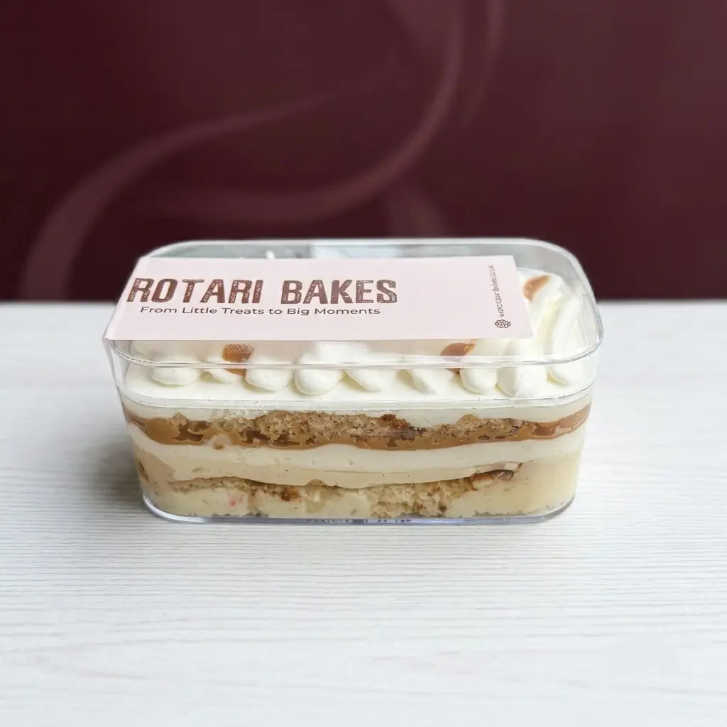 A rectangular clear plastic container holding a layered dessert cake with cream-colored frosting and cookie layers, topped with piped white frosting. A pink paper label on the lid reads 'ROTARI BAKES' with smaller text below, set on a white surface against a dark burgundy background.