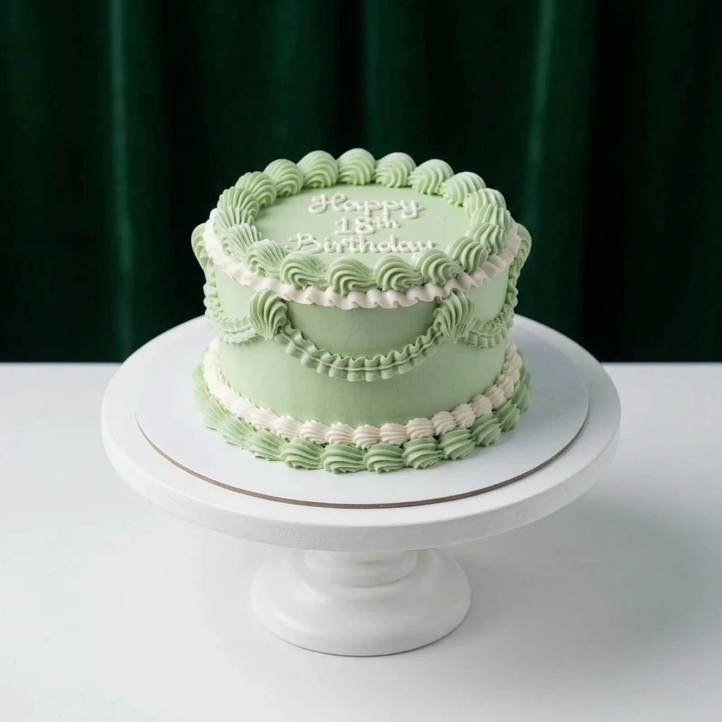 A round birthday cake with pale green frosting and piped decorative borders, placed on a white pedestal stand against a dark green curtain background. The top of the cake reads 'Happy 15th Birthday'.