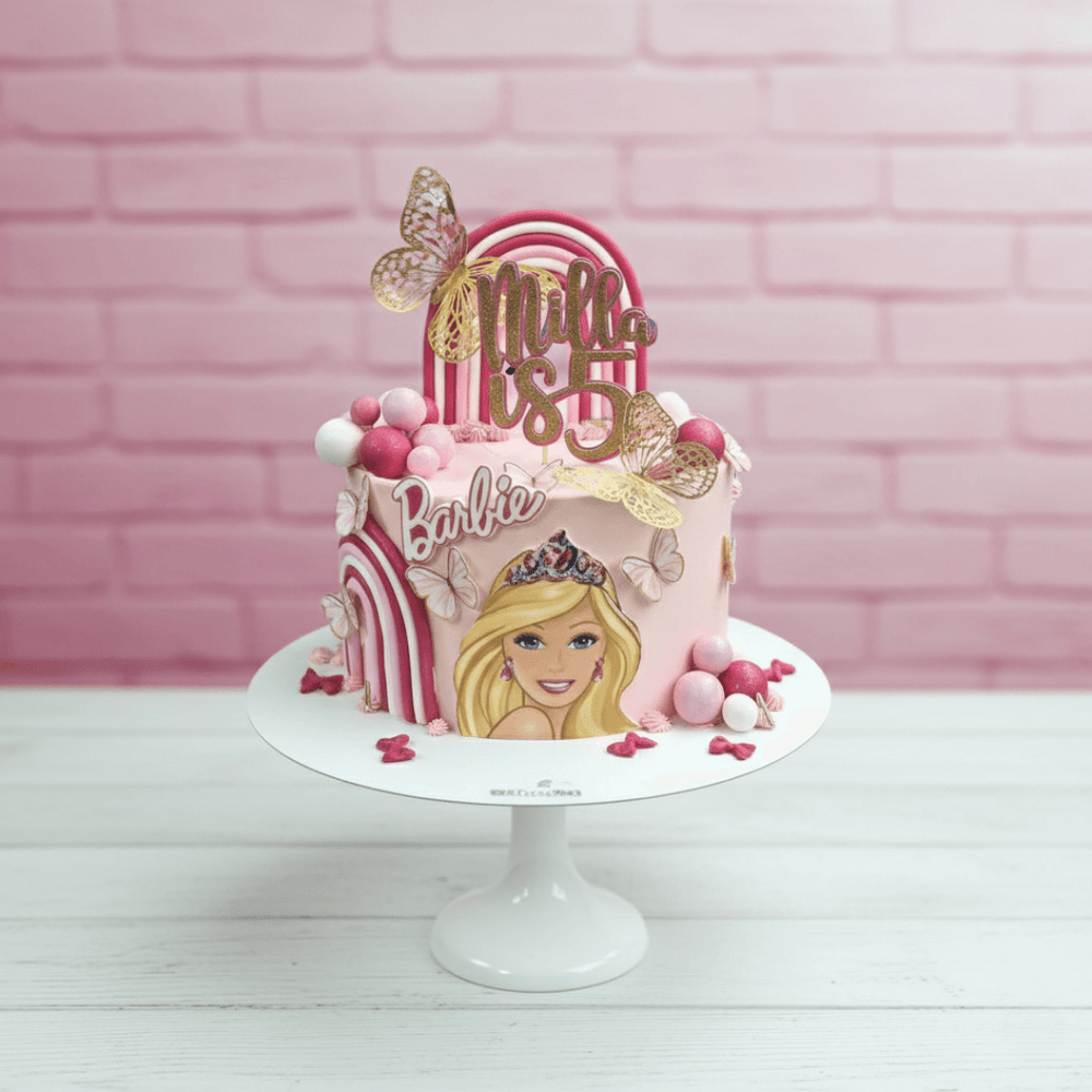 Barbie-themed birthday cake with pink and gold decorations on a white stand against a pink brick wall.
