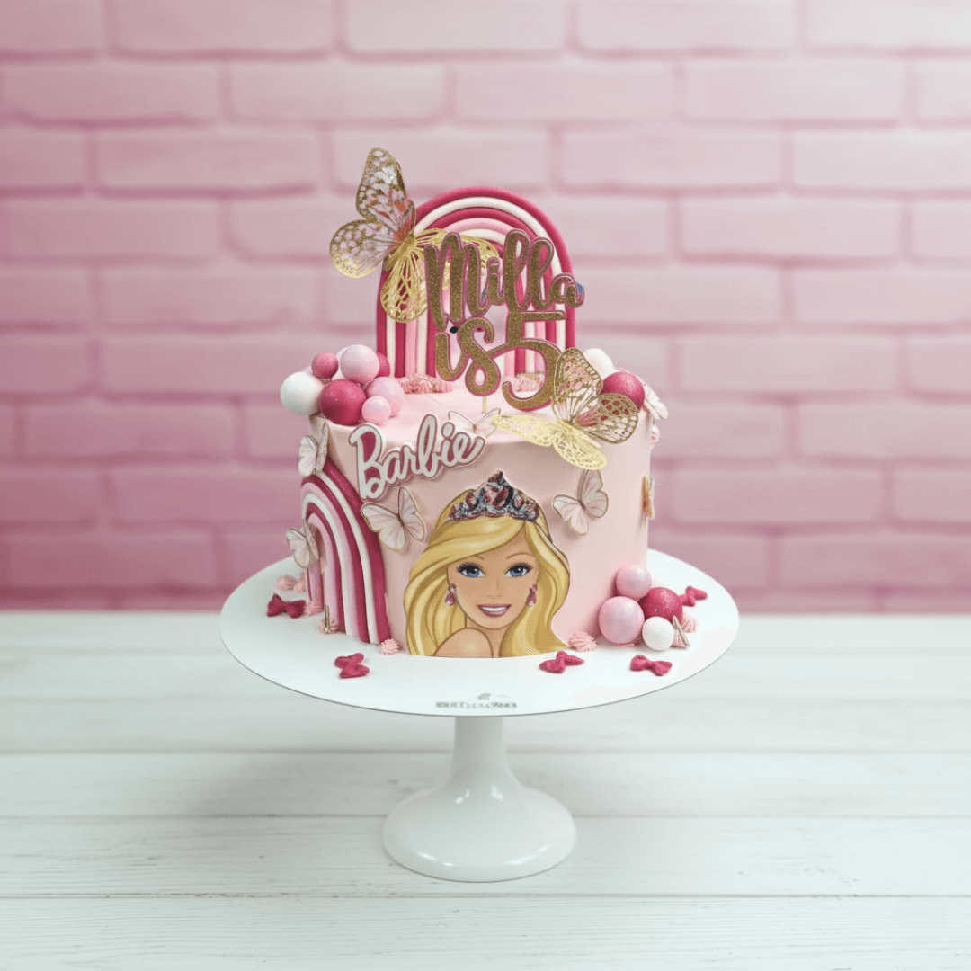 Barbie-themed birthday cake with pink and gold decorations on a white stand against a pink brick wall.
