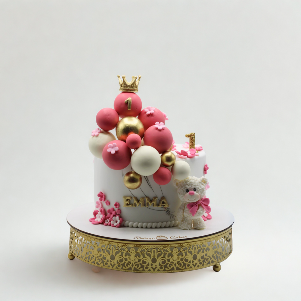 
                  
                    Small birthday cake with balloons and a crown on a decorative stand with a white background
                  
                