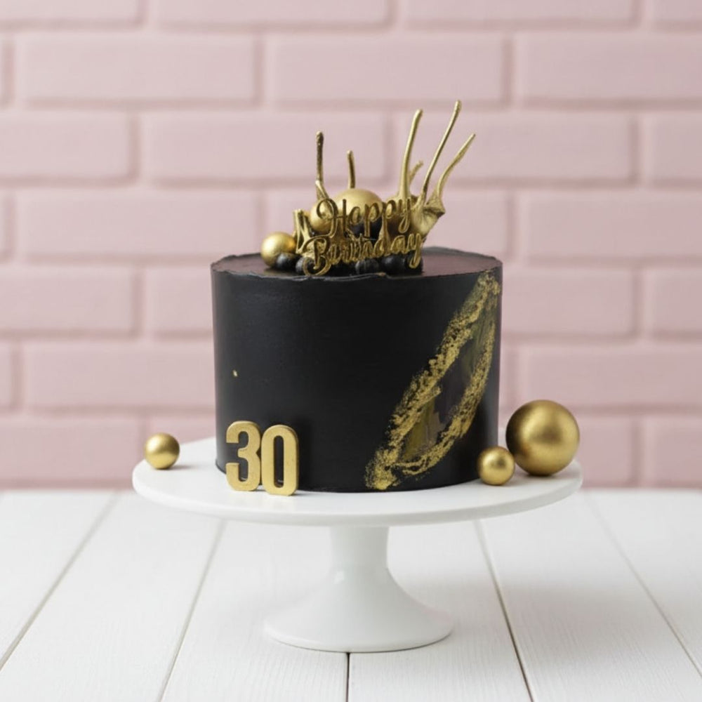 Black birthday cake with gold decorations on a white stand against a pink brick wall.