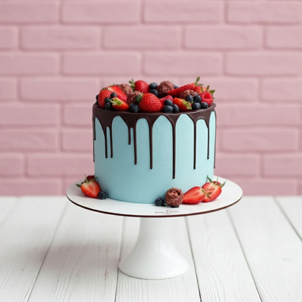 Light blue cake with chocolate drizzle and fresh berries on a white stand against a pink brick wall.