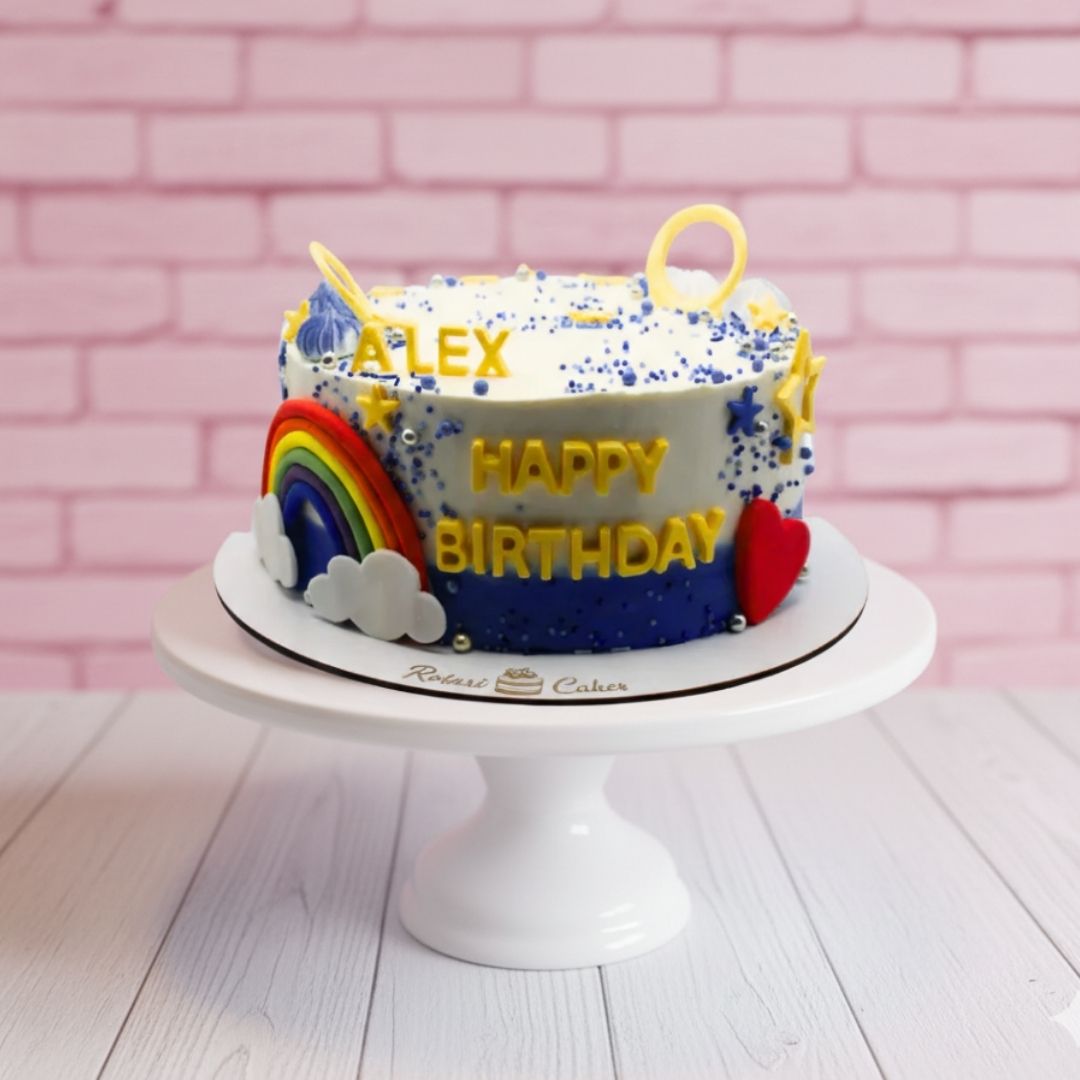 Colorful birthday cake with rainbow and stars on a white stand against a pink brick wall.