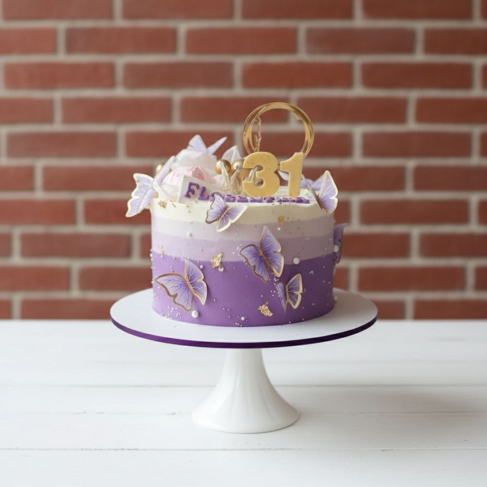 Purple birthday cake with butterfly decorations on a white stand against a brick wall.