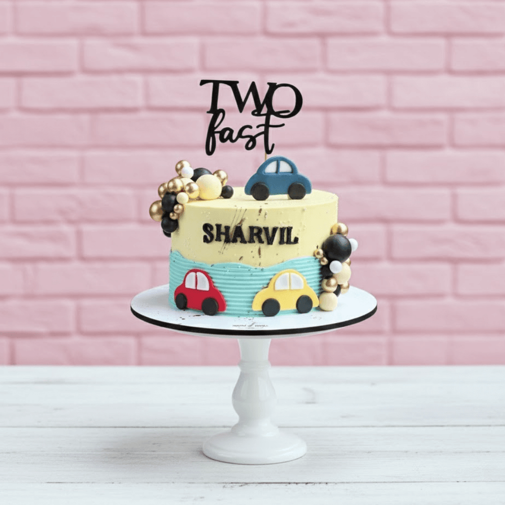 Children's birthday cake with colorful cars and 'Two Fast Sharvil' text against a pink brick wall.