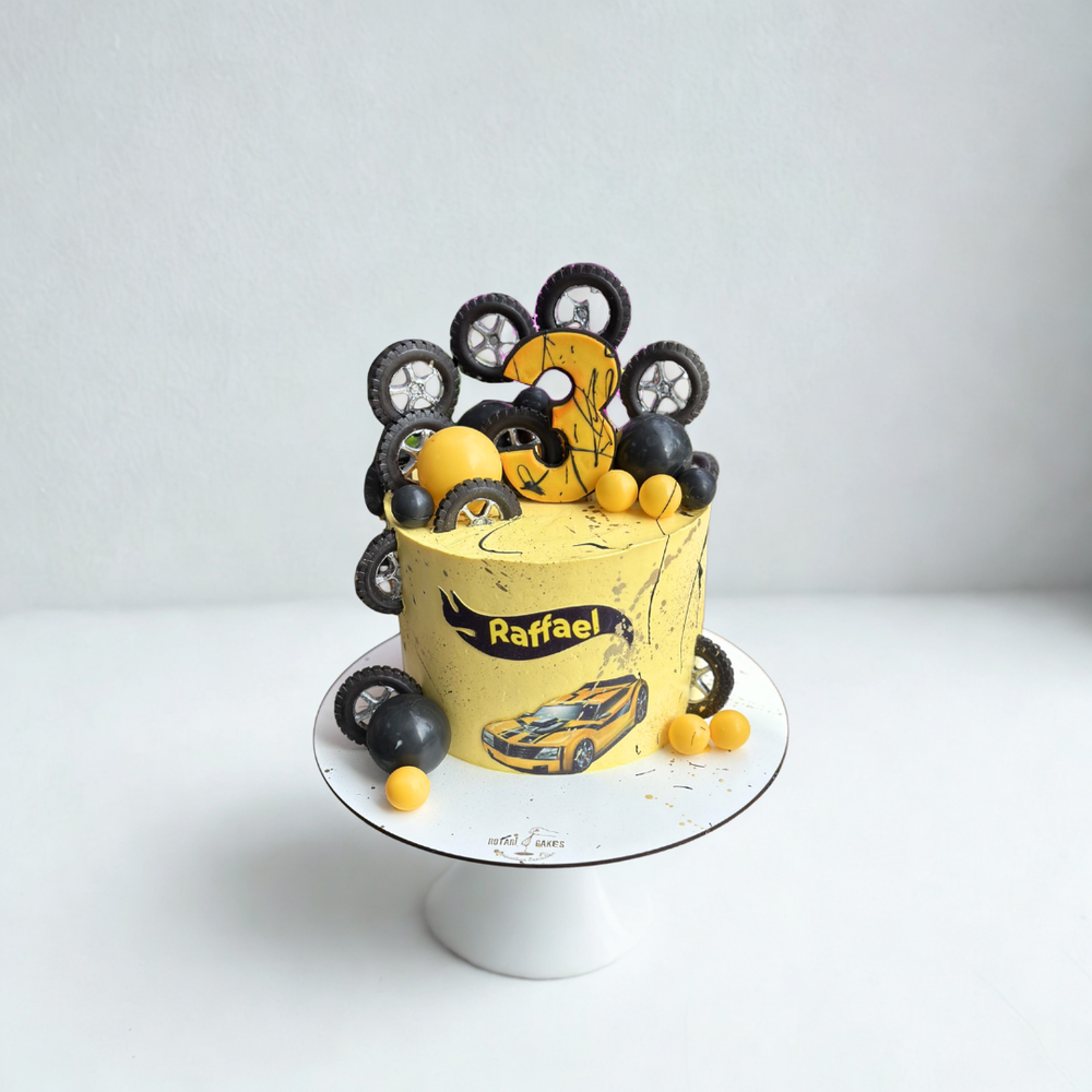 
                  
                    Yellow cake with car-themed decorations on a white stand against a light gray background
                  
                