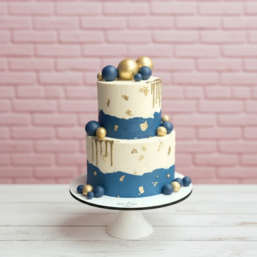 Two-tiered cake with blue and gold decorations on a white stand against a pink brick wall.
