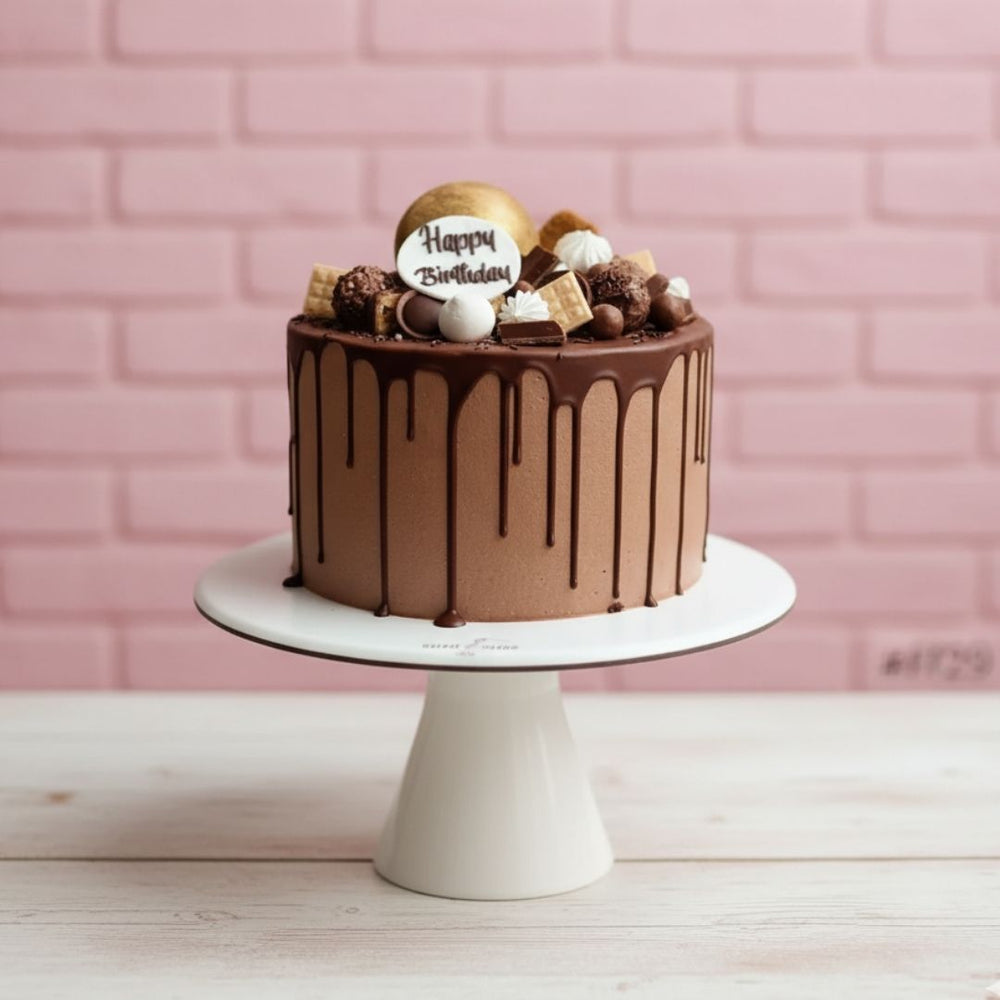 Chocolate cake with chocolate drizzle and decorative elements on a white cake stand against a pink brick wall.