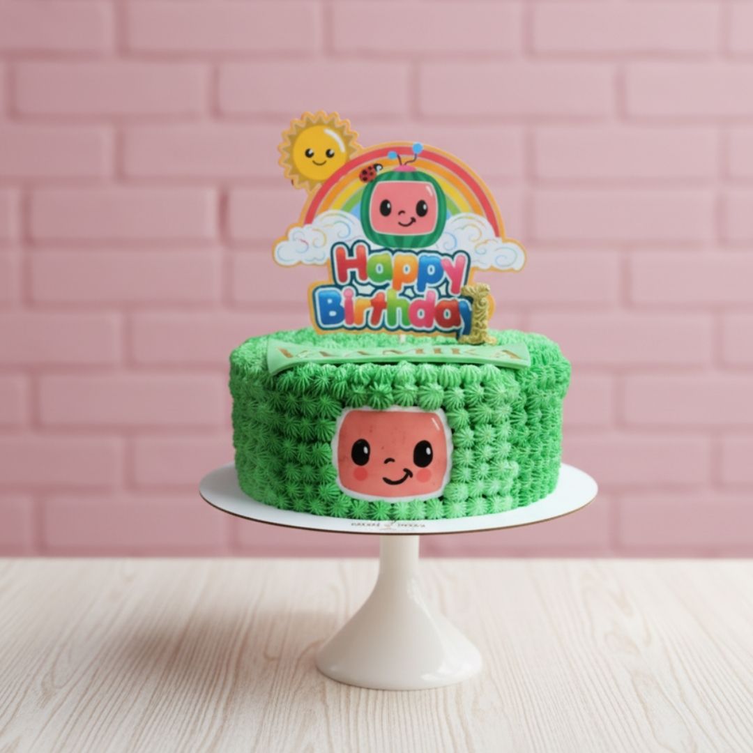 Green cocomelon birthday cake with a character-themed topper on a pink brick wall background