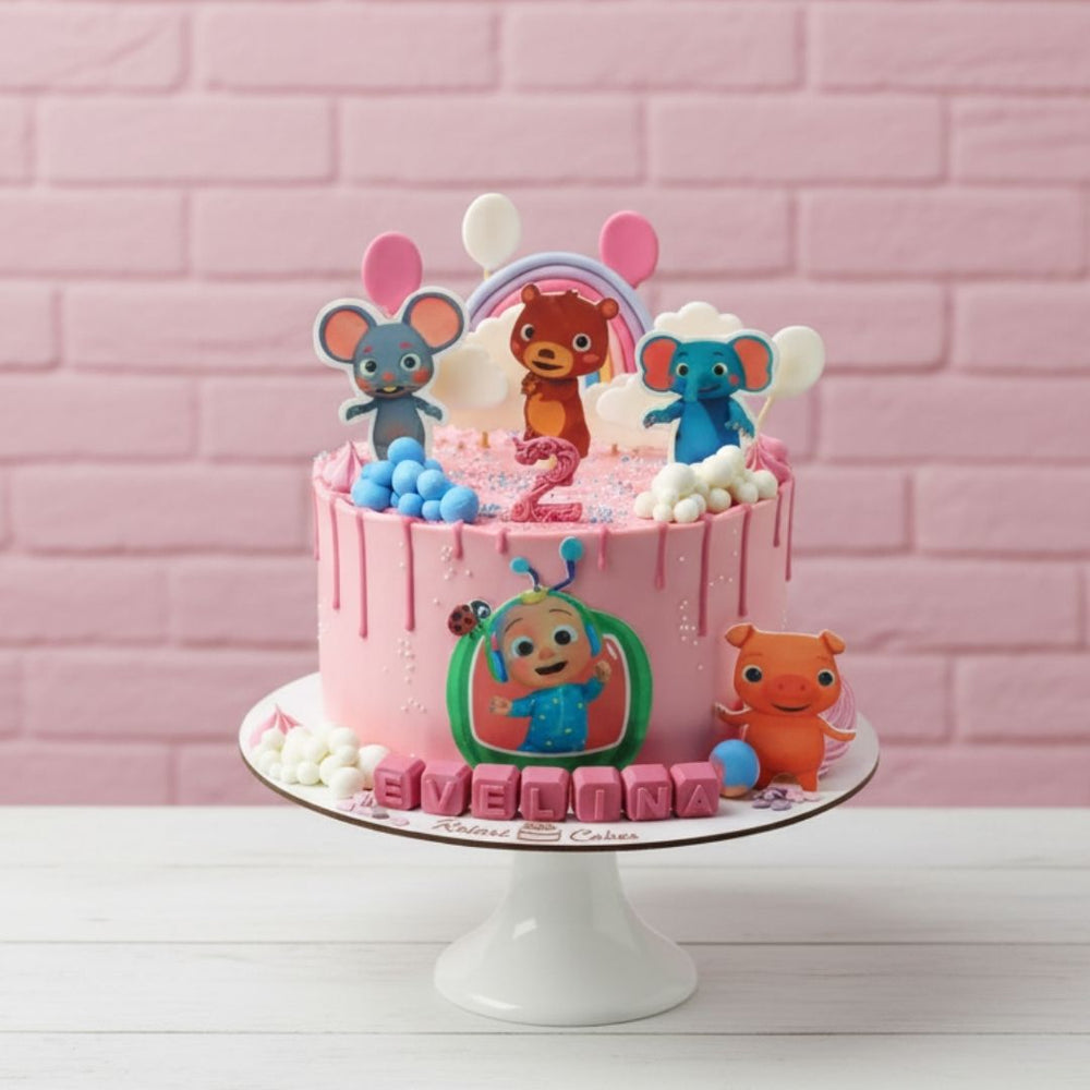 
                  
                    cocomelon theme Pink birthday cake with cartoon character toppers on a white stand against a pink brick wall.
                  
                