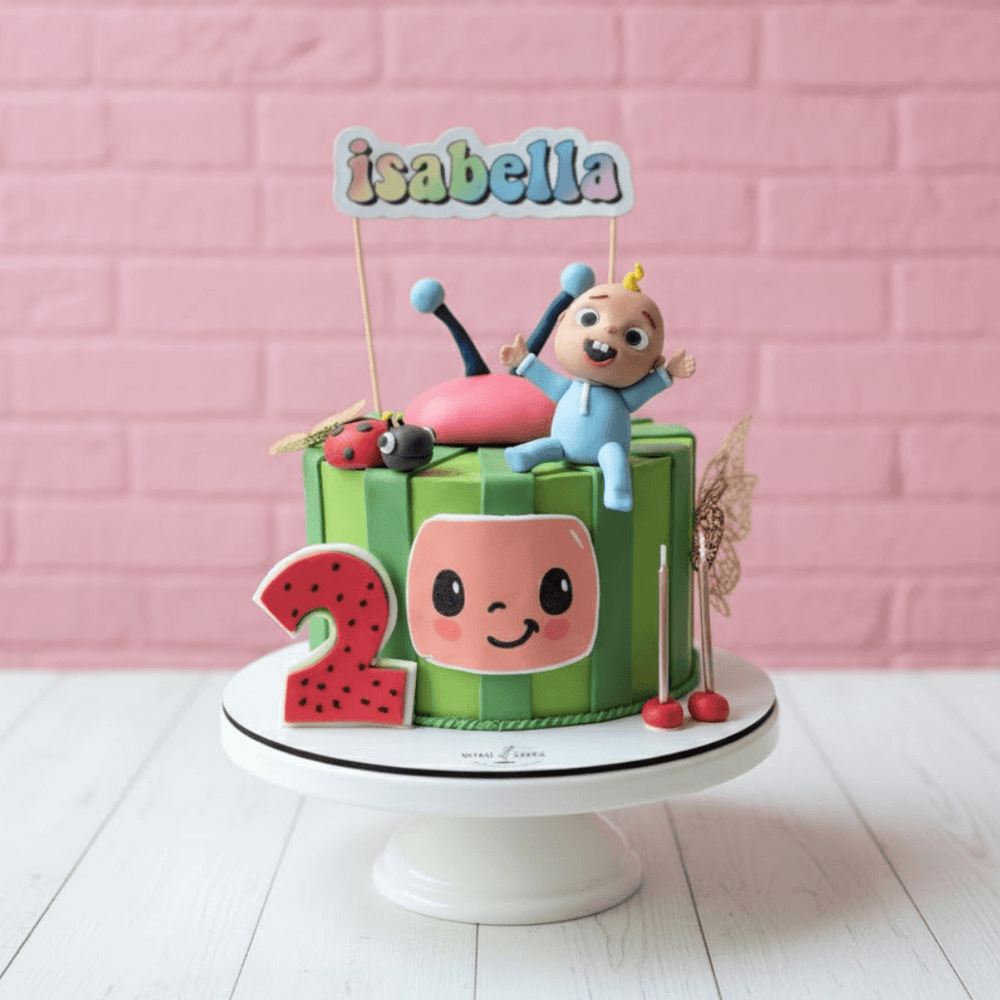 Cocomelon theme Children's birthday cake with cartoon character and 'isabella' topper against a pink brick wall.