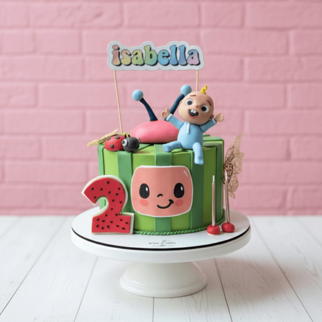 Cocomelon theme Children's birthday cake with cartoon character and 'isabella' topper against a pink brick wall.