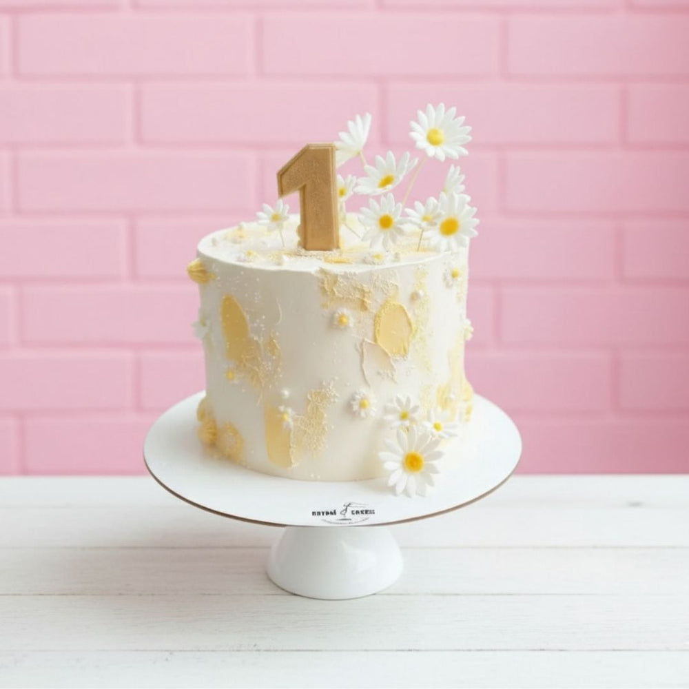 daisy theme Small white cake with yellow flowers on a white stand against a pink tiled wall.