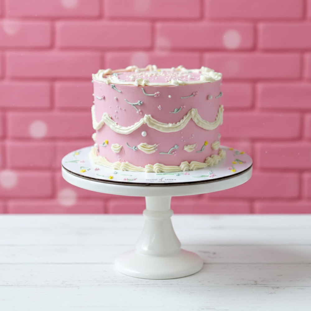 Pink flowers piping cake with white frosting on a white cake stand against a pink brick wall.
