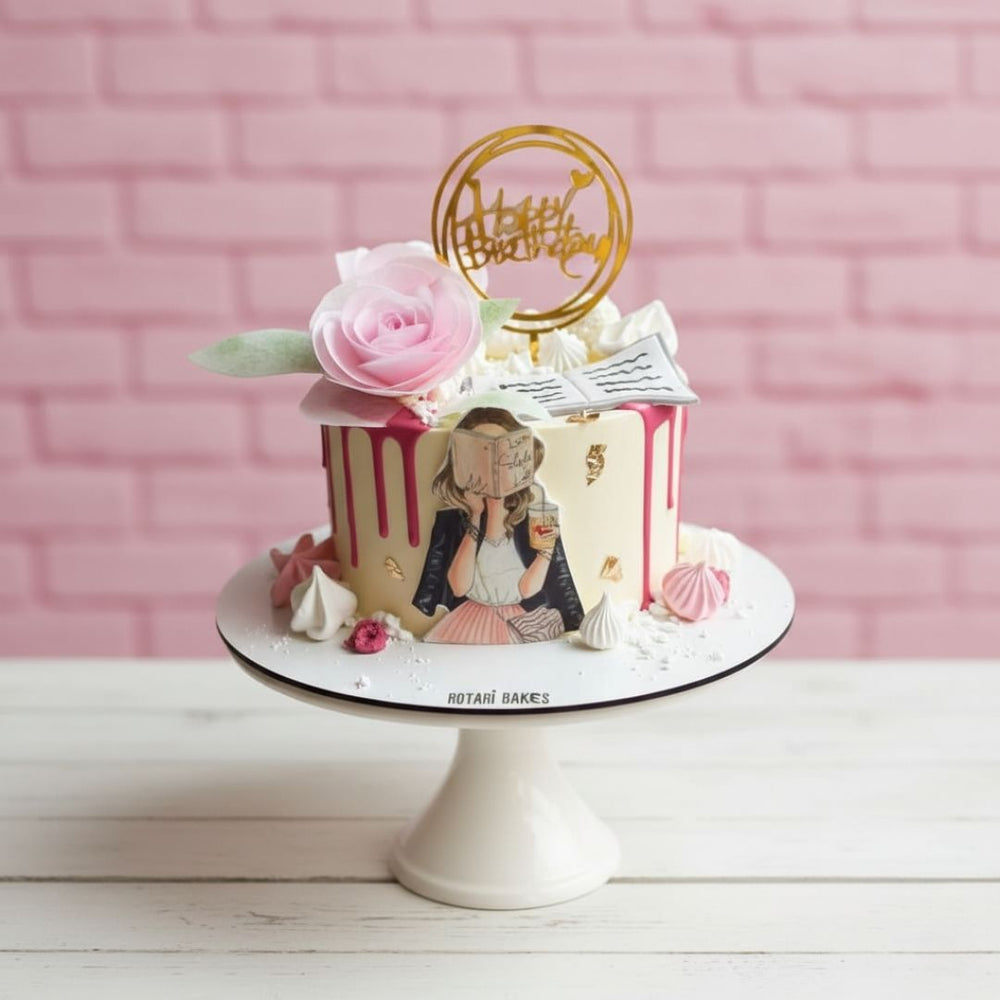 Decorative birthday cake with a pink rose and 'Happy Birthday' topper against a pink brick wall.
