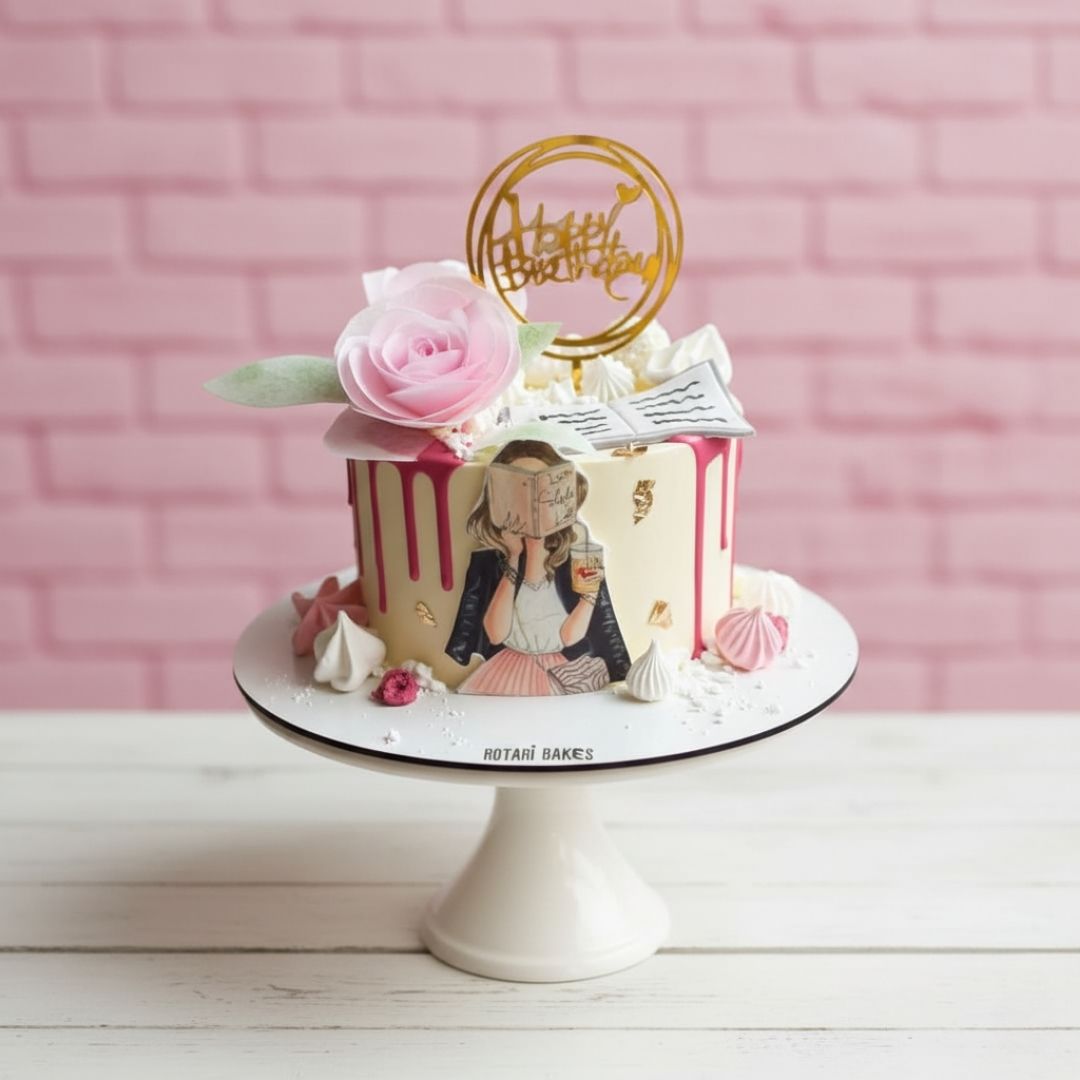 Decorative birthday cake with a pink rose and 'Happy Birthday' topper against a pink brick wall.