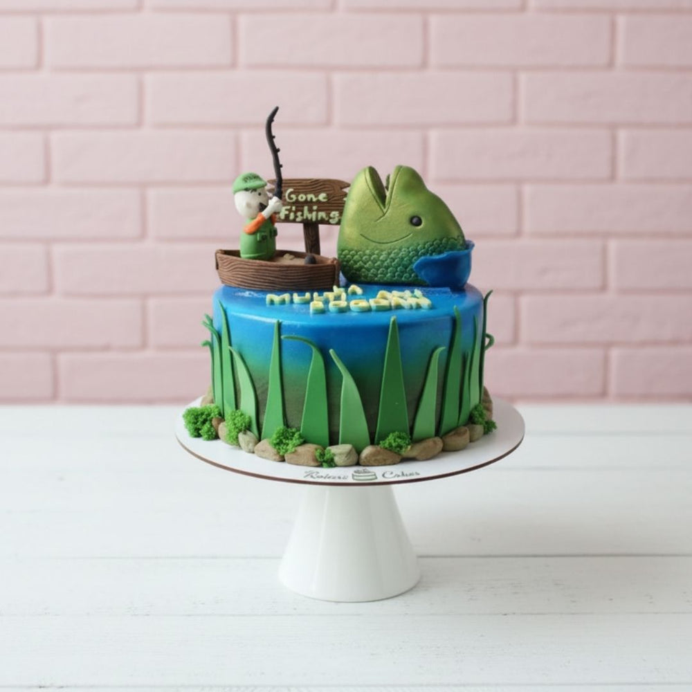 Fishing-themed cake with a fish and fishing rod decoration on a white stand against a pink brick wall.