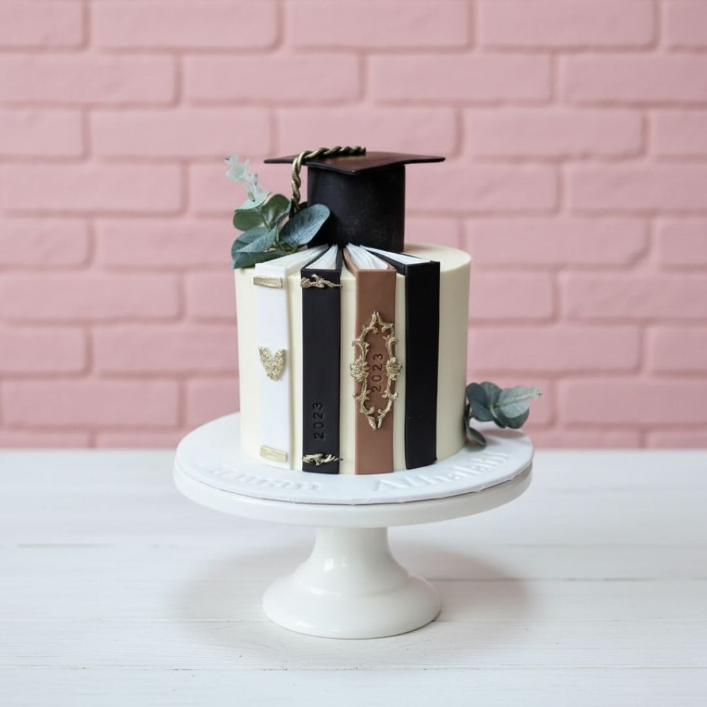 Graduation-themed cake with books and a cap on a white stand against a pink brick wall.