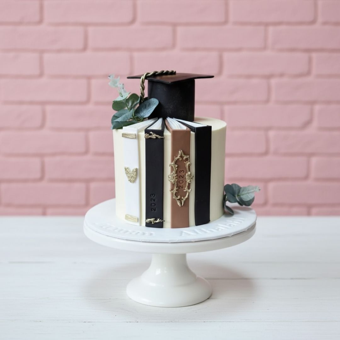 Graduation-themed cake with books and a cap on a white stand against a pink brick wall.