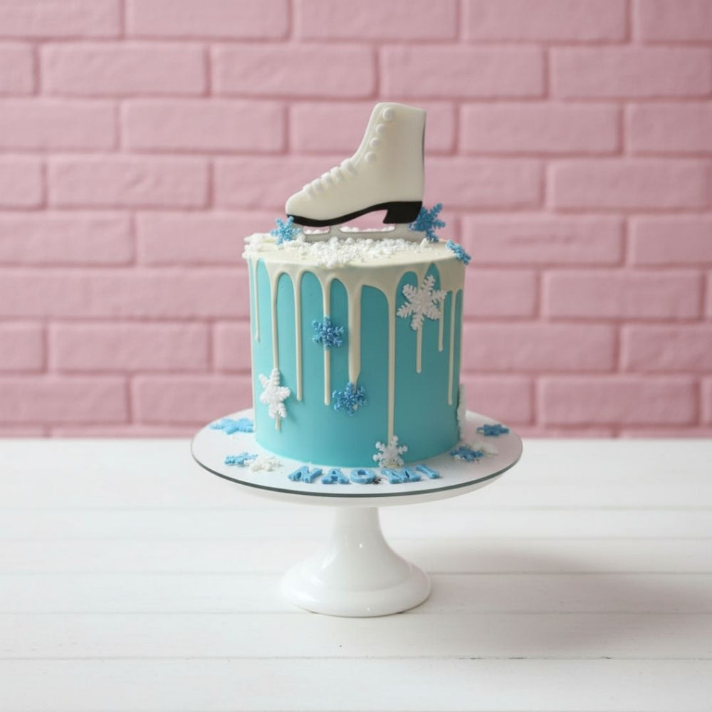 Teal cake with white icing, snowflakes, and an ice skate on a pink brick wall background
