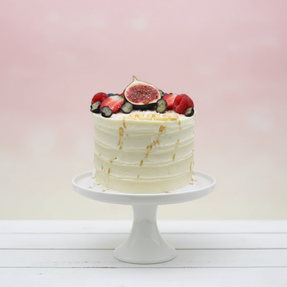 White cake with fruit toppings on a white stand against a light pink background