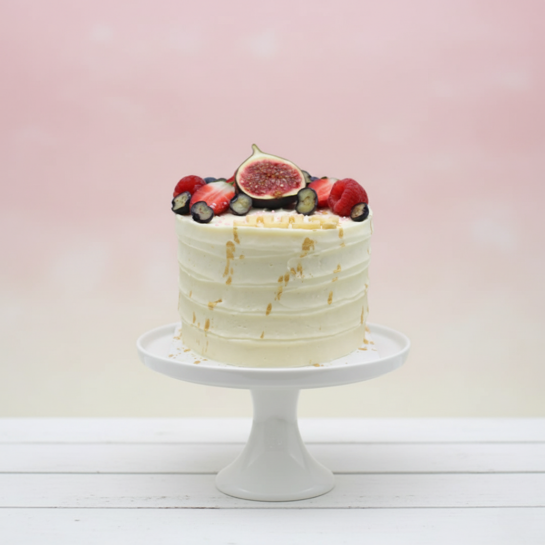 White cake with fruit toppings on a white stand against a light pink background
