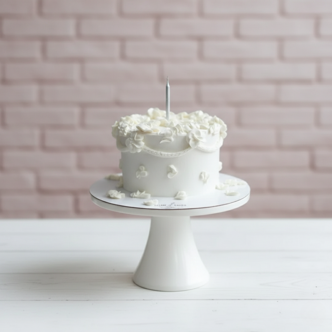White cake with a single candle on a white cake stand against a light pink brick wall.