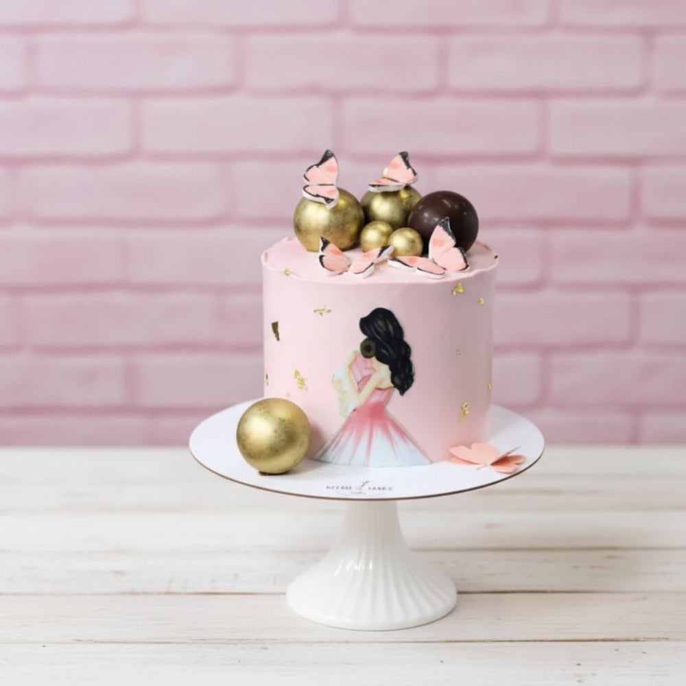 Decorative cake with pink frosting, gold balls, and butterfly toppers on a white stand against a pink brick wall.