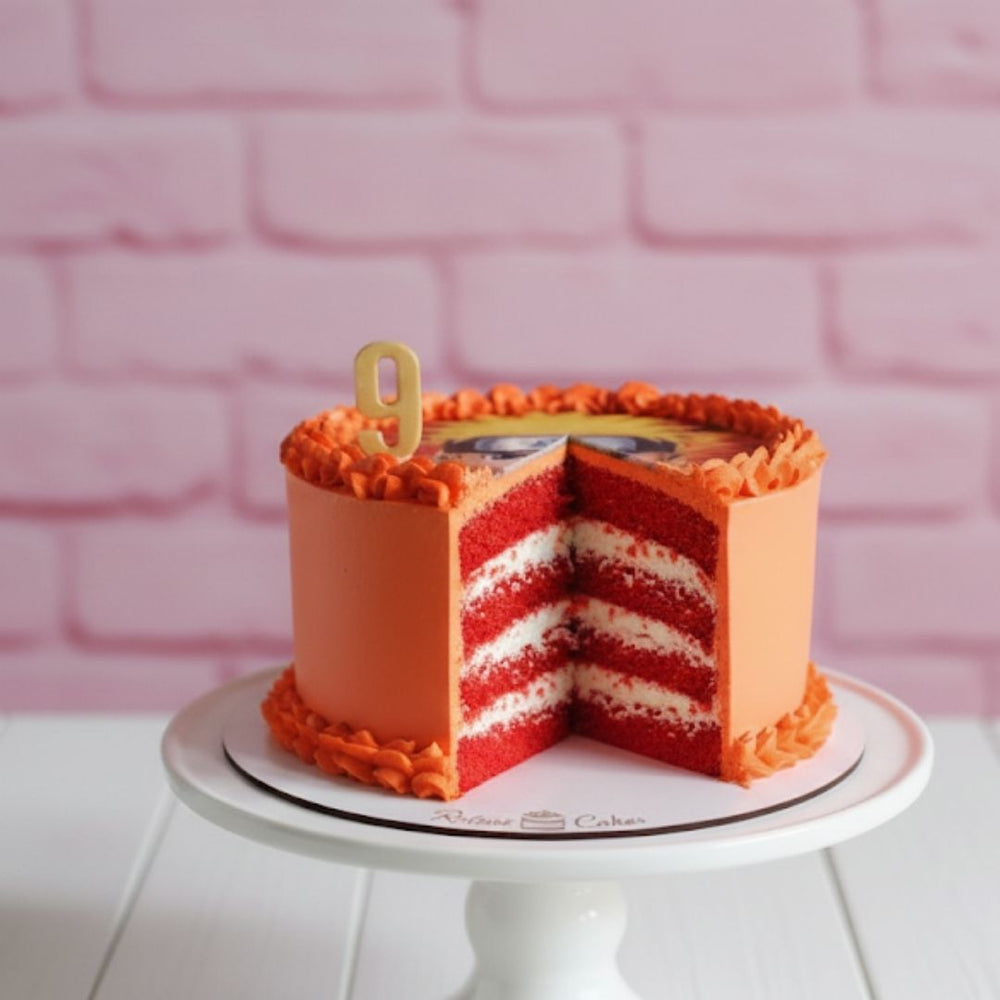 
                  
                    Red velvet flavour naruto theme orange cake with a slice cut out on a white stand against a pink brick wall.
                  
                