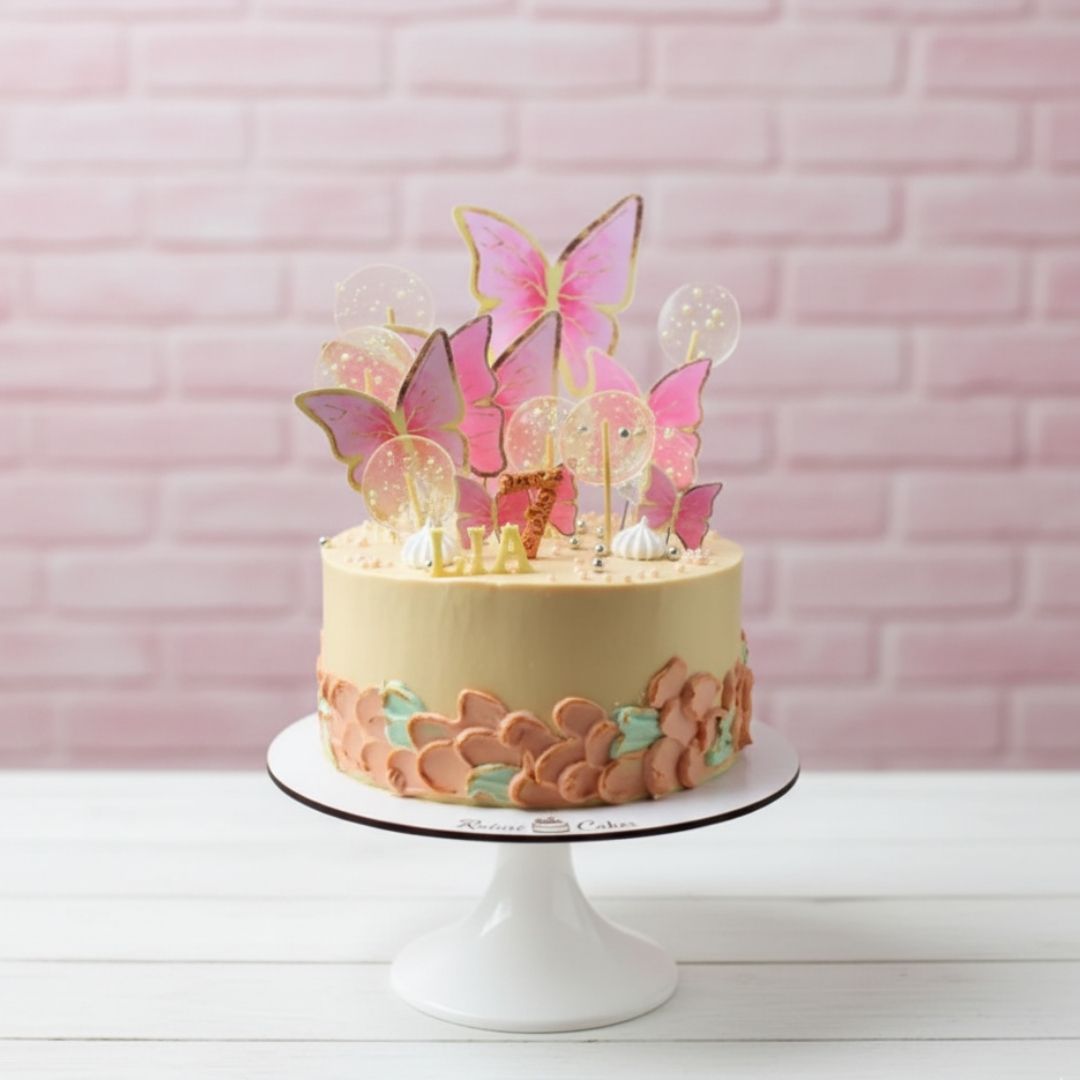 Decorative cake with butterfly toppers on a white stand against a pink brick wall.