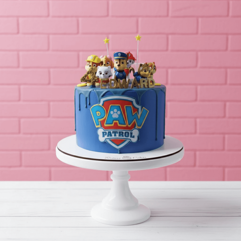 
                  
                    Paw Patrol birthday cake with character toppers on a white stand against a pink brick wall.
                  
                
