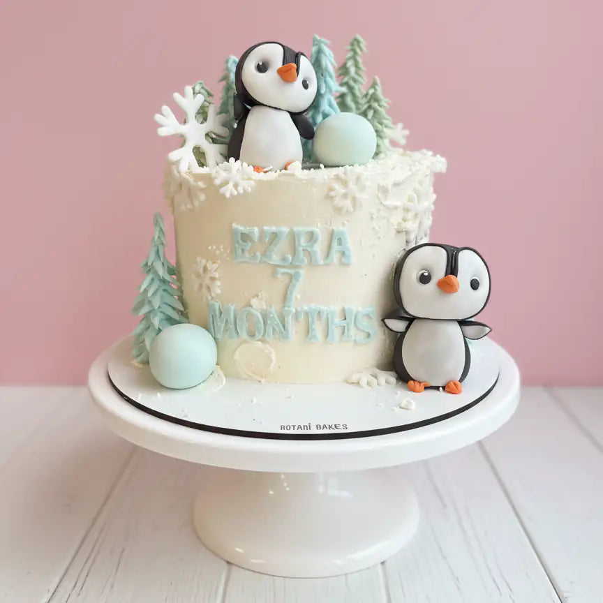 Cake with penguin decorations and 'Ezira 7 months' text on a white stand against a pink background