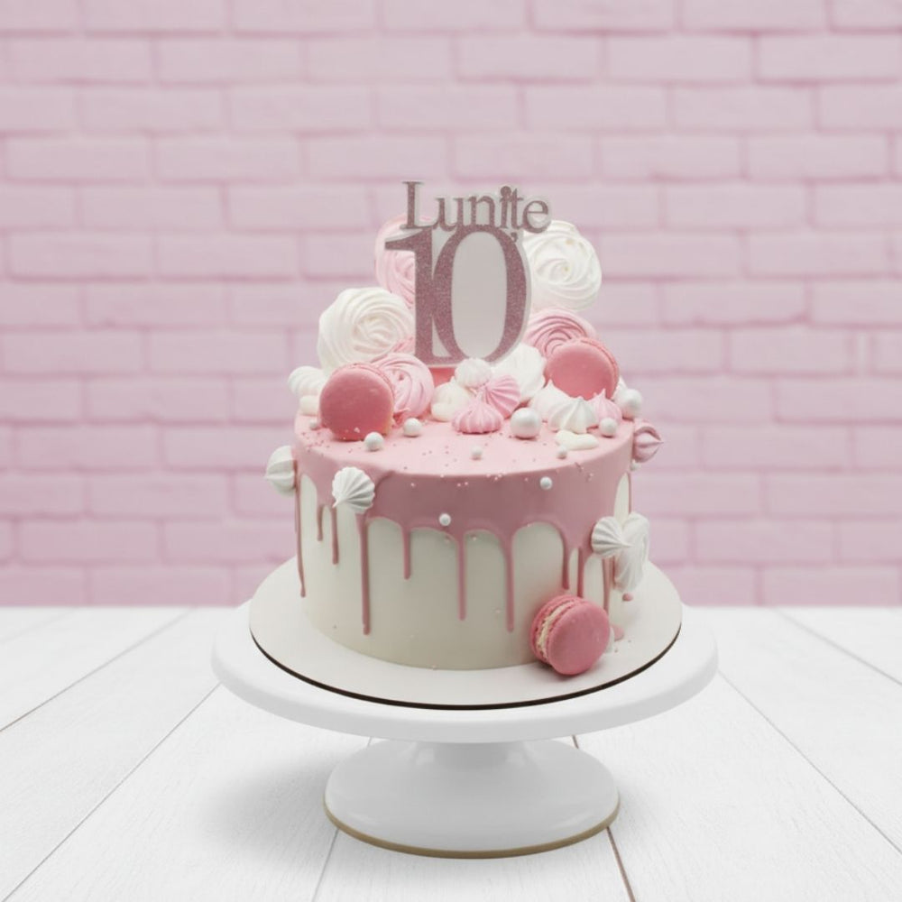 Pink birthday cake with decorative elements on a white stand against a pink brick wall.