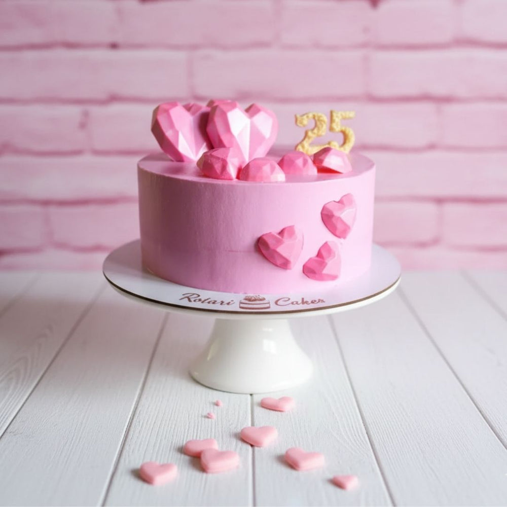 Pink birthday cake with heart decorations and a number '25' on a white stand against a pink brick wall.