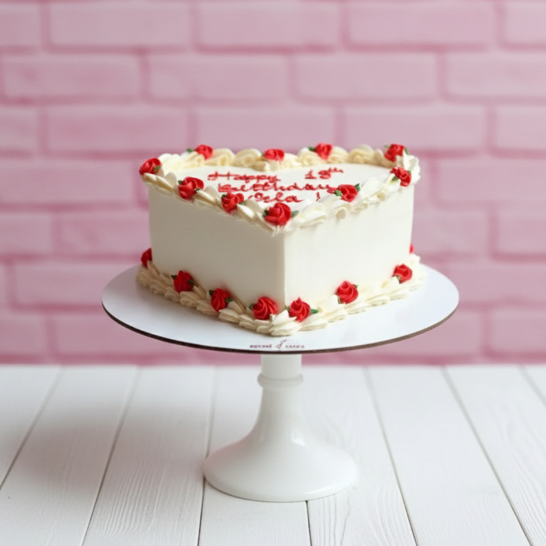 White heart shape birthday cake with red flowers on a white stand against a pink brick wall.