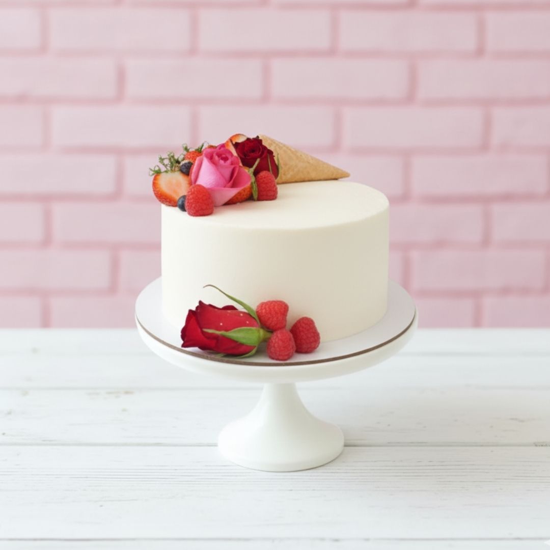 White cake with fruit and flowers on a white stand against a pink brick wall.