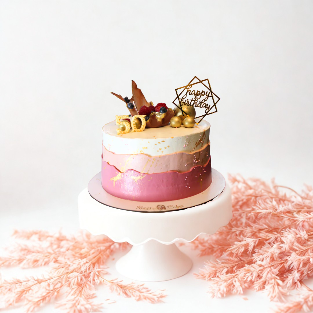 Pink and white birthday cake with gold accents on a white stand, surrounded by pink decorative elements.
